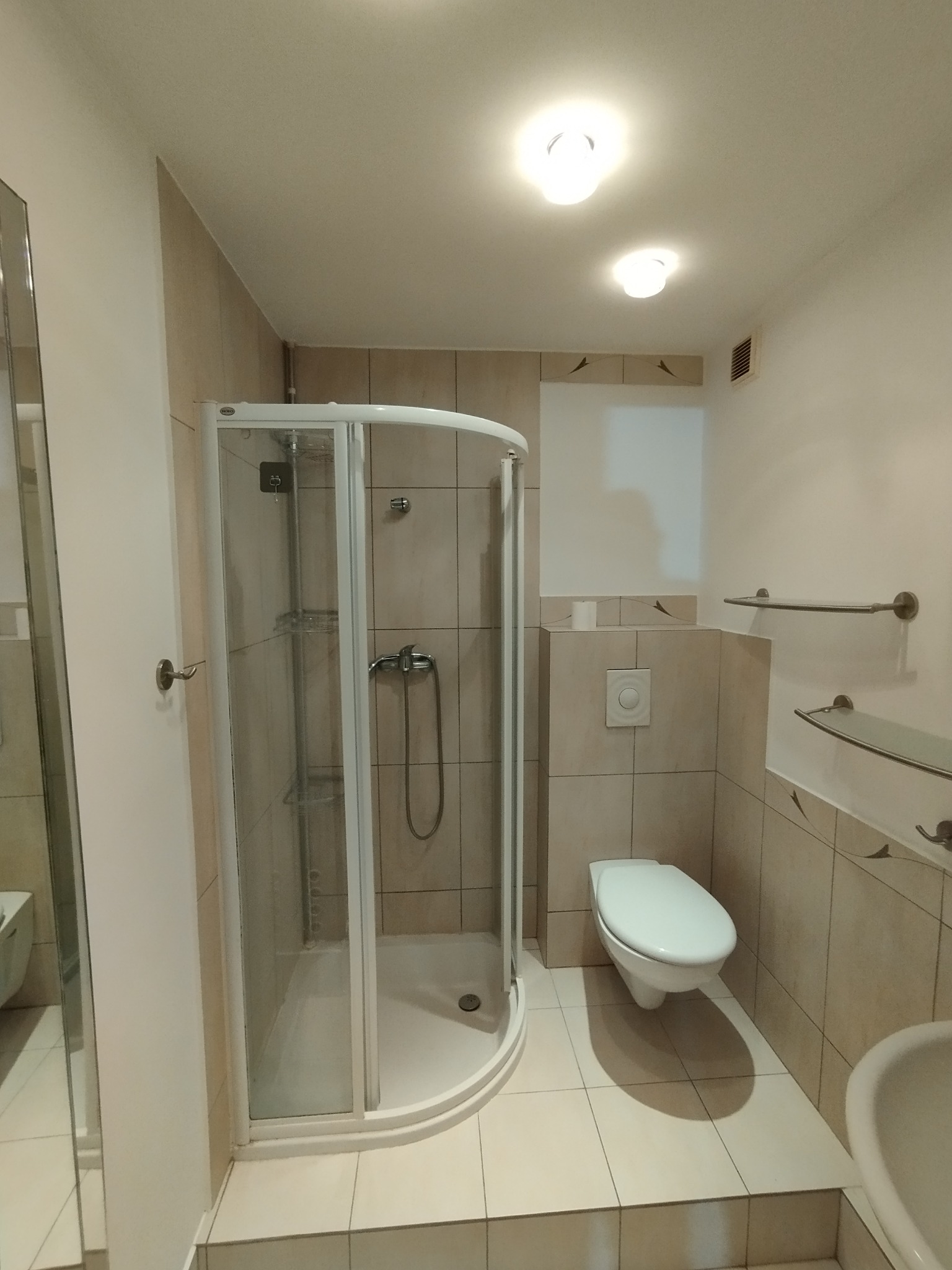 Jasna łazienka po remoncie: kabina prysznicowa z chromowaną armaturą, toaleta i umywalka. Beżowe płytki na ścianach i podłodze. Oświetlenie sufitowe.