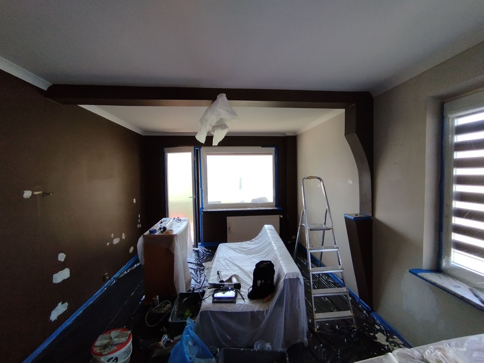 Pokój w trakcie malowania: ściany częściowo pokryte brązową farbą, zabezpieczone meble folią, drabina oparta o ścianę, widoczne ślady po szpachlowaniu.