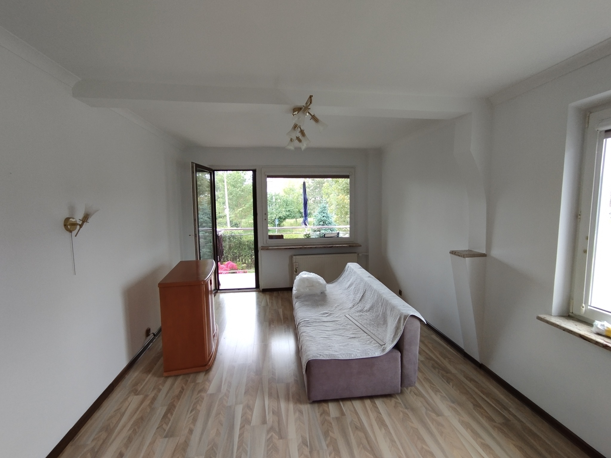 Jasne wnętrze pokoju po remoncie: świeżo pomalowane ściany, drewniana podłoga, okno z widokiem na zieleń. Sofa przykryta folią ochronną, gotowa do użytku.