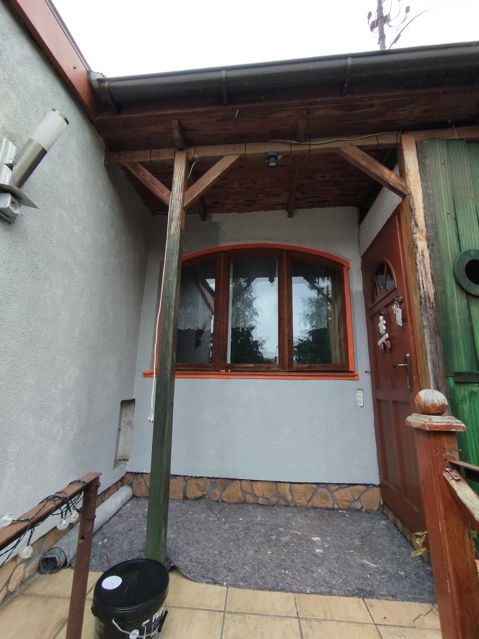 Szara elewacja domu z drewnianą werandą i oknem z pomarańczową ramą. Widoczny fragment drzwi i wiadro z farbą. Oświetlenie zewnętrzne i dekoracje.