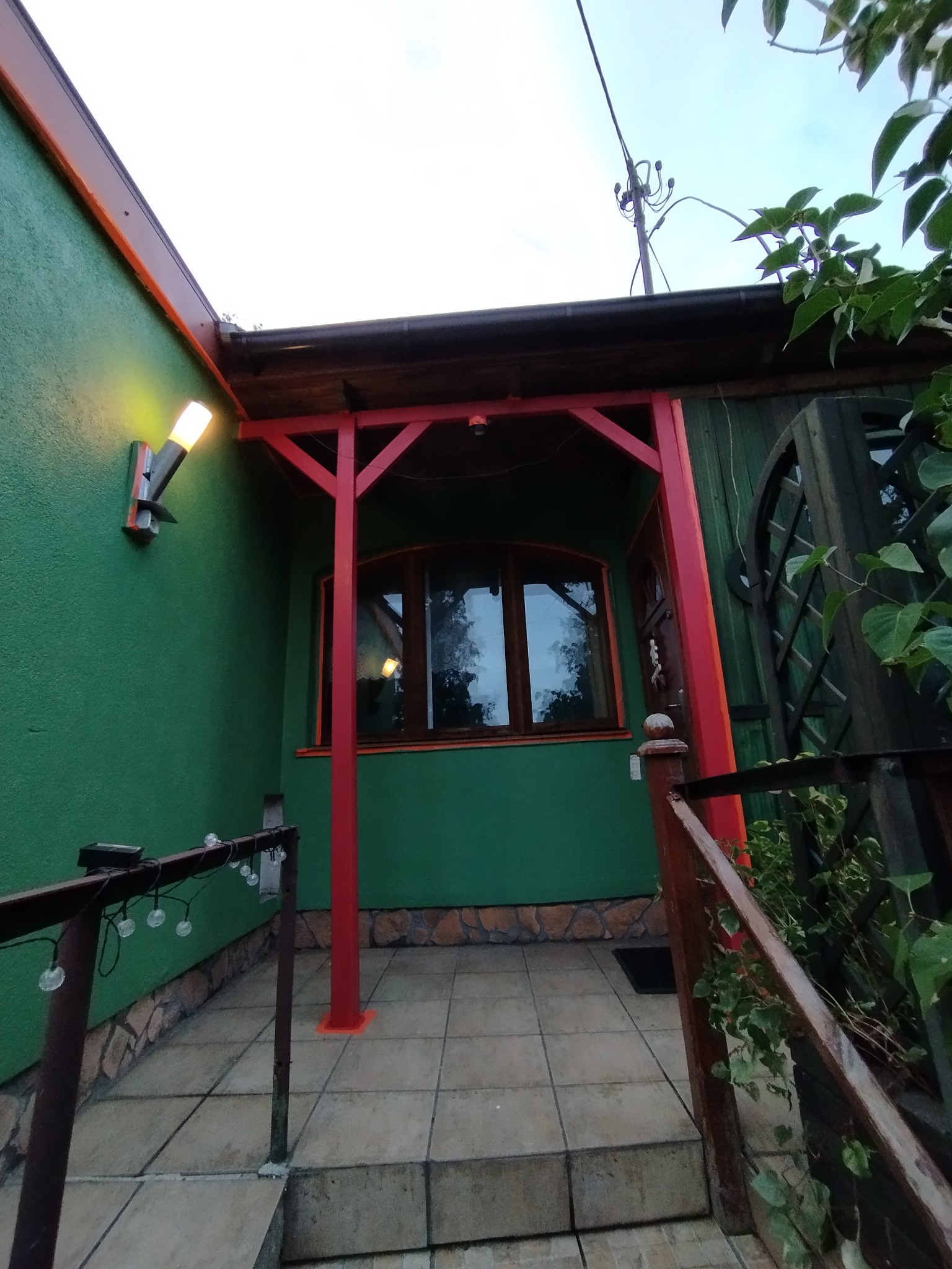 Zielony dom z czerwonymi elementami konstrukcyjnymi i oświetleniem zewnętrznym. Widoczne okno z drewnianą ramą i fragment tarasu z balustradą.