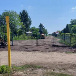 Ż&oacute;łta koparko-ładowarka CAT podczas niwelacji terenu pod montaż ogrodzenia, w tle widoczny walec drogowy i fragmenty siatki ogrodzeniowej.