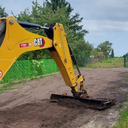 ErdeLux - Żółta ramię koparki CAT z równiarką wyrównuje ziemię w Zawierciu, widoczny fragment zielonego ogrodzenia i drzewa w tle.