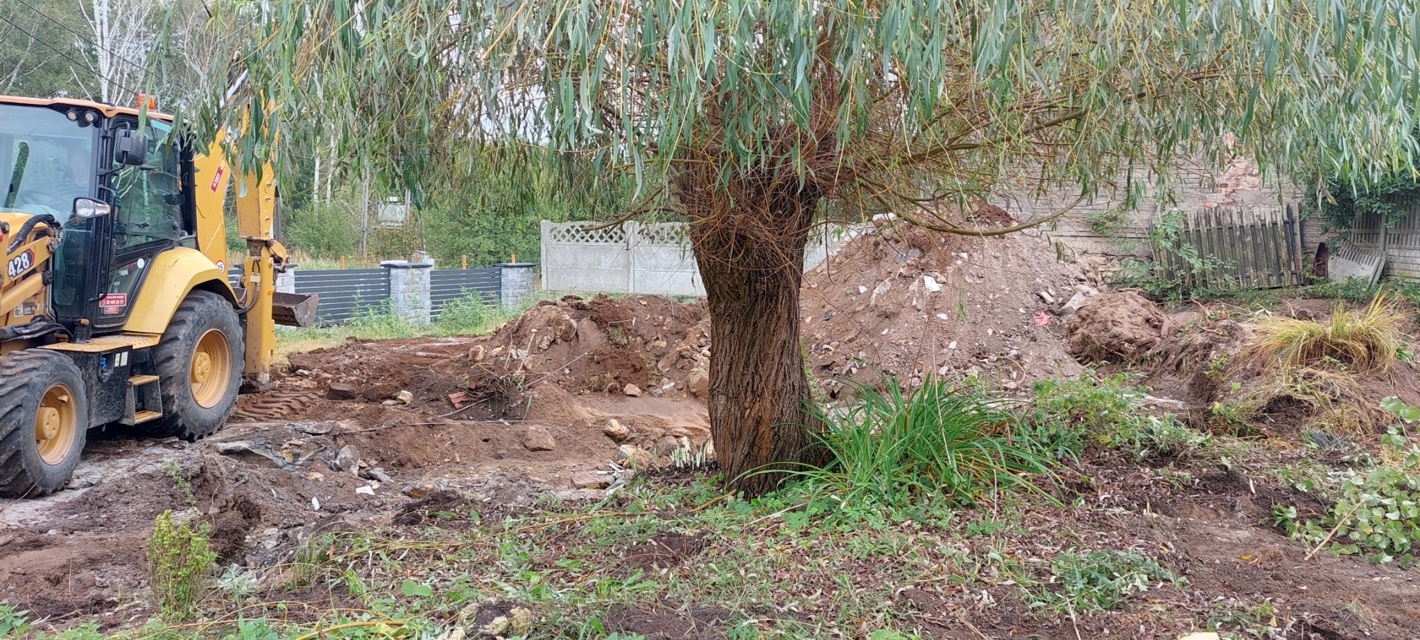 Żółta koparko-ładowarka na placu budowy, widoczny wykop ziemi, hałda gruzu i drzewo z długimi gałęziami.