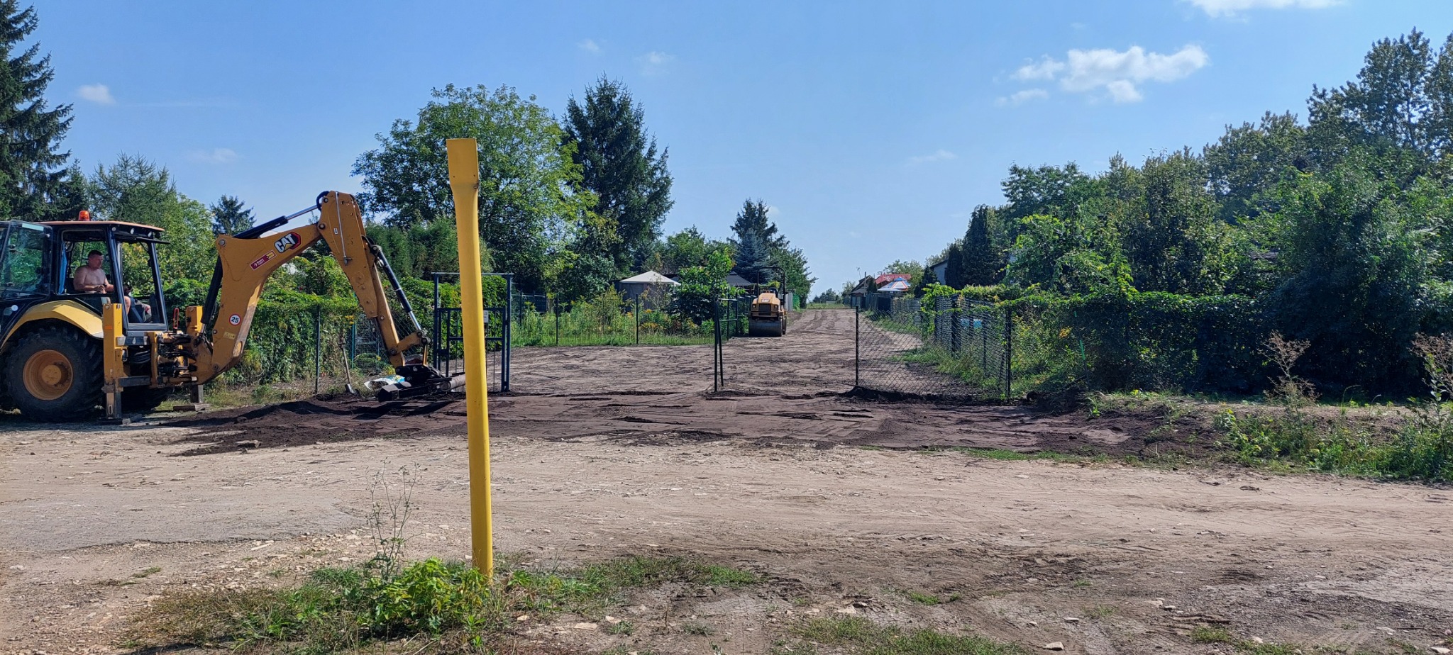 Żółta koparko-ładowarka CAT podczas niwelacji terenu pod montaż ogrodzenia, w tle widoczny walec drogowy i fragmenty siatki ogrodzeniowej.