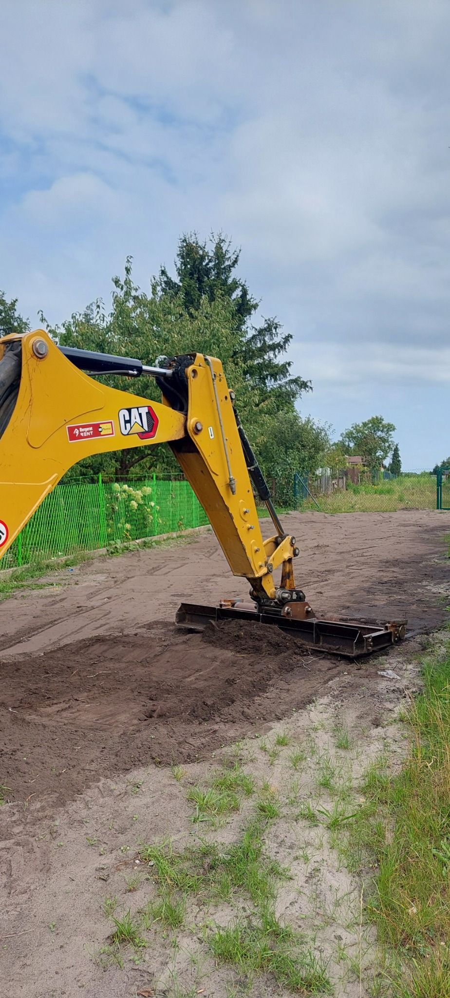 Żółta ramię koparki CAT z równiarką wyrównuje ziemię w Zawierciu, widoczny fragment zielonego ogrodzenia i drzewa w tle.