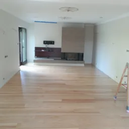 Jasne, drewniane podłogi po cyklinowaniu w przestronnym, odświeżonym salonie z kominkiem i przygotowanymi ścianami do dalszych prac wykończeniowych; widoczna drabina malarska.