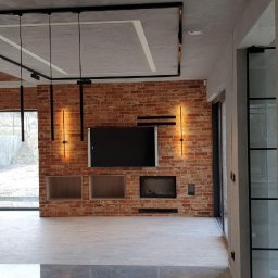 Usługi budowlane Grzegorz Ornawka - Salon z ceglaną ścianą, minimalistycznym oświetleniem i dużymi oknami, widok z korytarza na przestronne wnętrze z elementami dekoracyjnymi w stylu industrialnym.