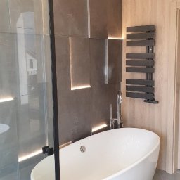 Usługi budowlane Grzegorz Ornawka - Nowoczesna łazienka z wolnostojącą białą wanną, szklaną kabiną prysznicową z czarnymi profilami, dekoracyjną ścianą z podświetlanych paneli w odcieniach szarości oraz drewnianą ścianą z grzejnikiem...