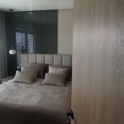 Usługi budowlane Grzegorz Ornawka - Częściowy widok sypialni z łóżkiem w neutralnych kolorach, widoczny przez otwarte drzwi z drewnianą okleiną, w tle okno z żaluzjami.