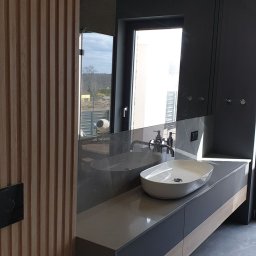 Usługi budowlane Grzegorz Ornawka - Nowoczesna łazienka z umywalką nablatową, szarą armaturą i drewnianymi panelami ściennymi, widok na zewnątrz przez okno odbijający mikser betoniarski.