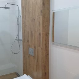 Usługi budowlane Grzegorz Ornawka - Nowoczesna łazienka z podwieszaną miską WC, kabiną prysznicową typu walk-in i ścianą wyłożoną płytkami imitującymi drewno, lustro z podświetleniem.