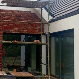 Usługi budowlane Grzegorz Ornawka - Fragment elewacji budynku w trakcie wykańczania z czerwoną cegłą klinkierową, widoczne rusztowanie z narzędziami i resztkami cegieł, okna z ciemnymi ramami i fragment dachu z dachówką.