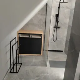 Nowoczesna łazienka na poddaszu z szarymi płytkami imitującymi marmur, czarną armaturą prysznicową, szafką z jasnego drewna i czarnymi frontami oraz czarnym, minimalistycznym wieszakiem na ręczniki.