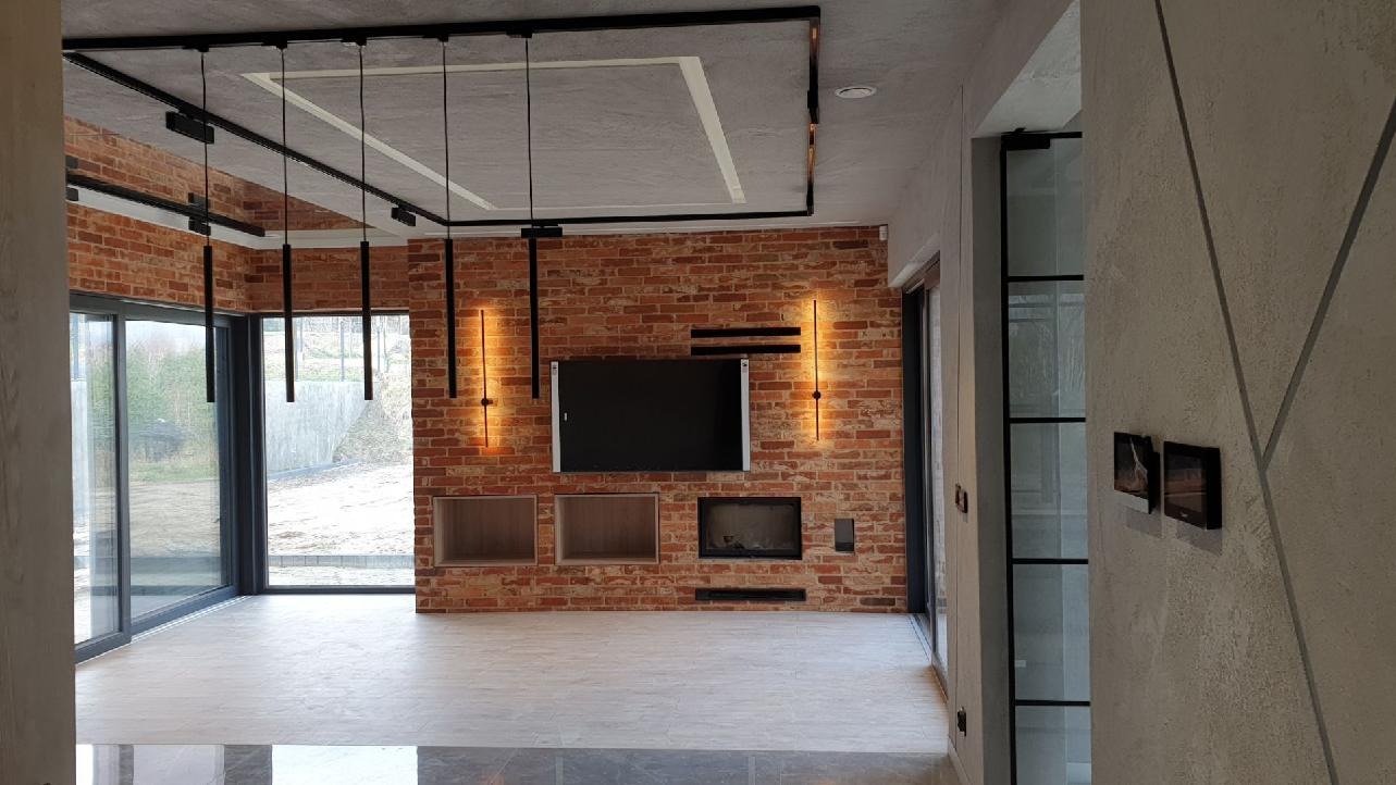 Salon z ceglaną ścianą, minimalistycznym oświetleniem i dużymi oknami, widok z korytarza na przestronne wnętrze z elementami dekoracyjnymi w stylu industrialnym.