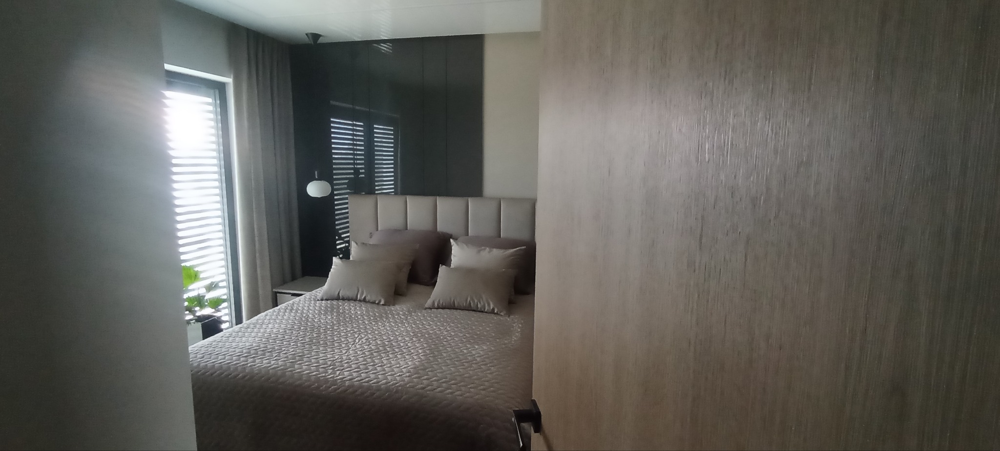 Częściowy widok sypialni z łóżkiem w neutralnych kolorach, widoczny przez otwarte drzwi z drewnianą okleiną, w tle okno z żaluzjami.