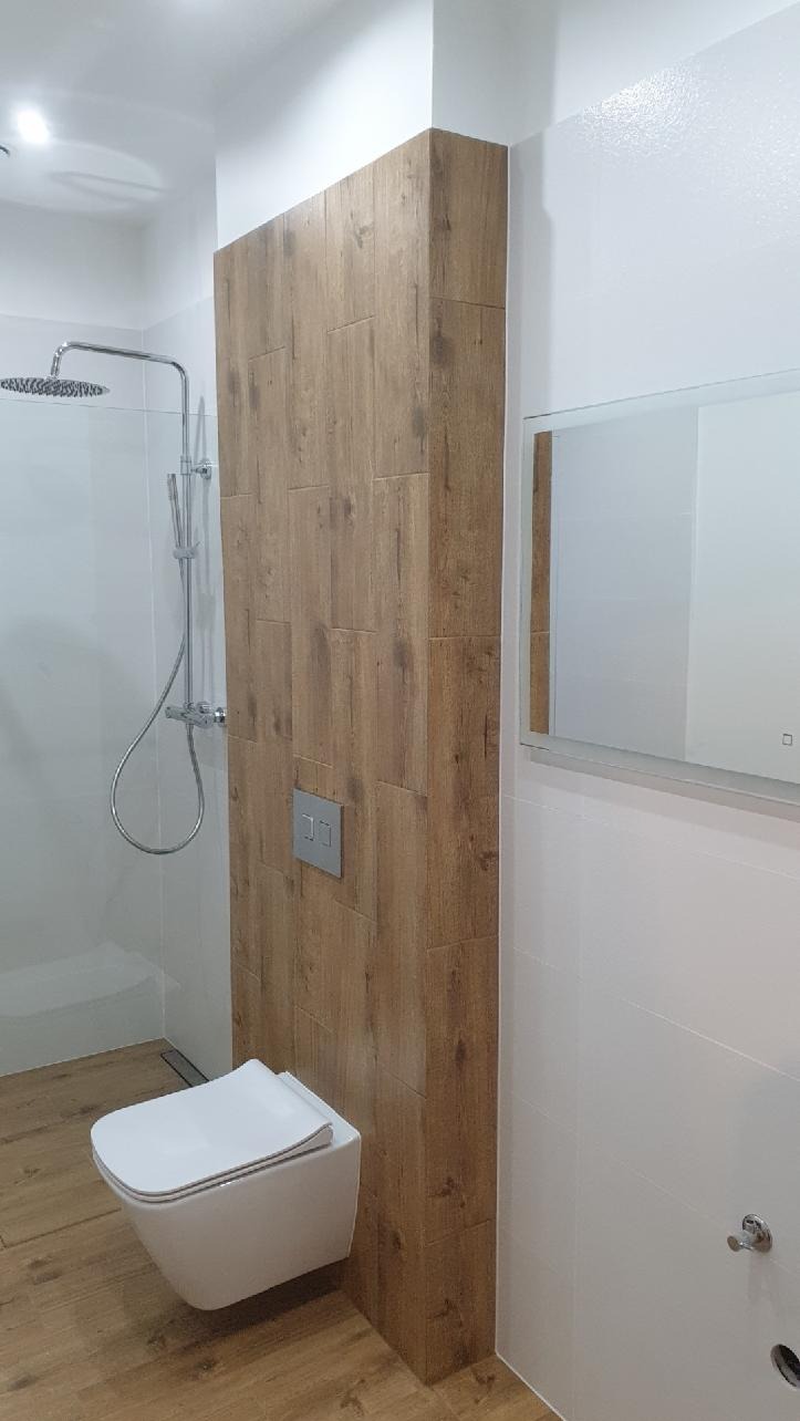 Nowoczesna łazienka z podwieszaną miską WC, kabiną prysznicową typu walk-in i ścianą wyłożoną płytkami imitującymi drewno, lustro z podświetleniem.