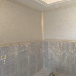 IDO&JAN SPÓŁKA Z OGRANICZONĄ ODPOWIEDZIALNOŚCIĄ - Wykończona łazienka z szarymi płytkami imitującymi marmur na dole ścian i beżową, teksturowaną farbą na górze, oddzielone ozdobną listwą. Widoczny fragment sufitu z oświetleniem.