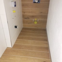 IDO&JAN SPÓŁKA Z OGRANICZONĄ ODPOWIEDZIALNOŚCIĄ - Przygotowanie do montażu podtynkowego stelaża WC na ścianie wyłożonej panelami drewnopodobnymi z widocznymi przyłączami, białe płytki na sąsiedniej ścianie, podłoga wyłożona panelami imitującymi...