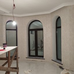 IDO&JAN SPÓŁKA Z OGRANICZONĄ ODPOWIEDZIALNOŚCIĄ - Pokój w trakcie prac wykończeniowych, z widocznymi trzema oknami w kształcie łuków, pomalowanymi ścianami z delikatnym wzorem, rozstawionym rusztowaniem malarskim i wiszącą żarówką.