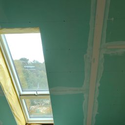 IDO&JAN SPÓŁKA Z OGRANICZONĄ ODPOWIEDZIALNOŚCIĄ - Montaż płyt gipsowo-kartonowych na poddaszu wokół okna dachowego, zabezpieczonego folią ochronną, widoczne szpachlowane łączenia i pracownik na drabinie.