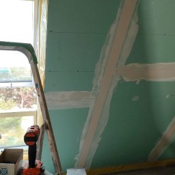 IDO&JAN SPÓŁKA Z OGRANICZONĄ ODPOWIEDZIALNOŚCIĄ - Poddasze w trakcie zabudowy płytami gipsowo-kartonowymi, widoczne łączenia płyt wzmocnione taśmą i zaszpachlowane, drabina oparta o okno dachowe, wiertarko-wkrętarka i karton z narzędziami obok.