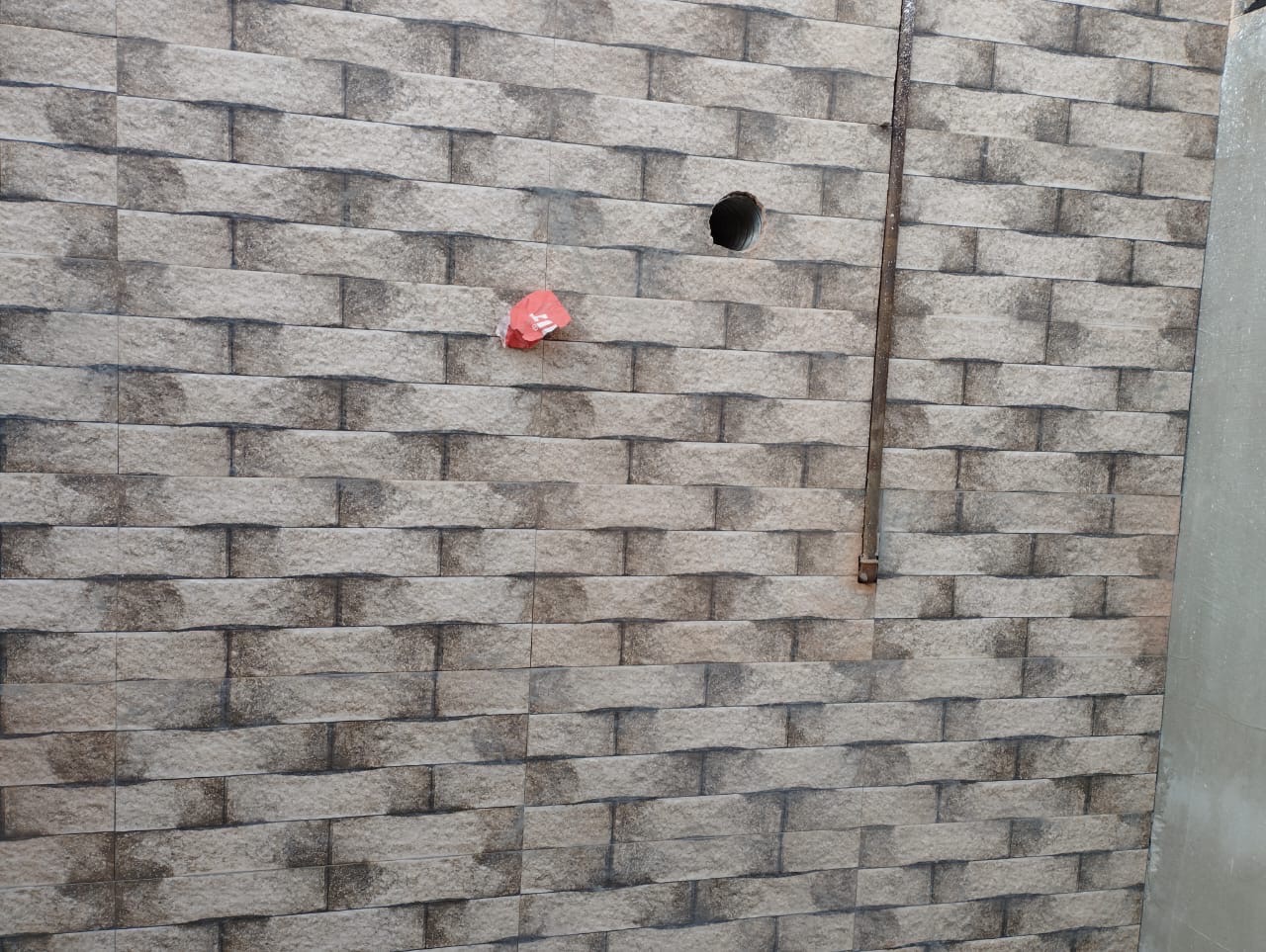 Ściana w trakcie układania płytek imitujących cegłę w Warszawie, widoczny otwór na instalację i pionowa rurka. Na jednej z płytek przyklejony mały kawałek czerwonej taśmy.