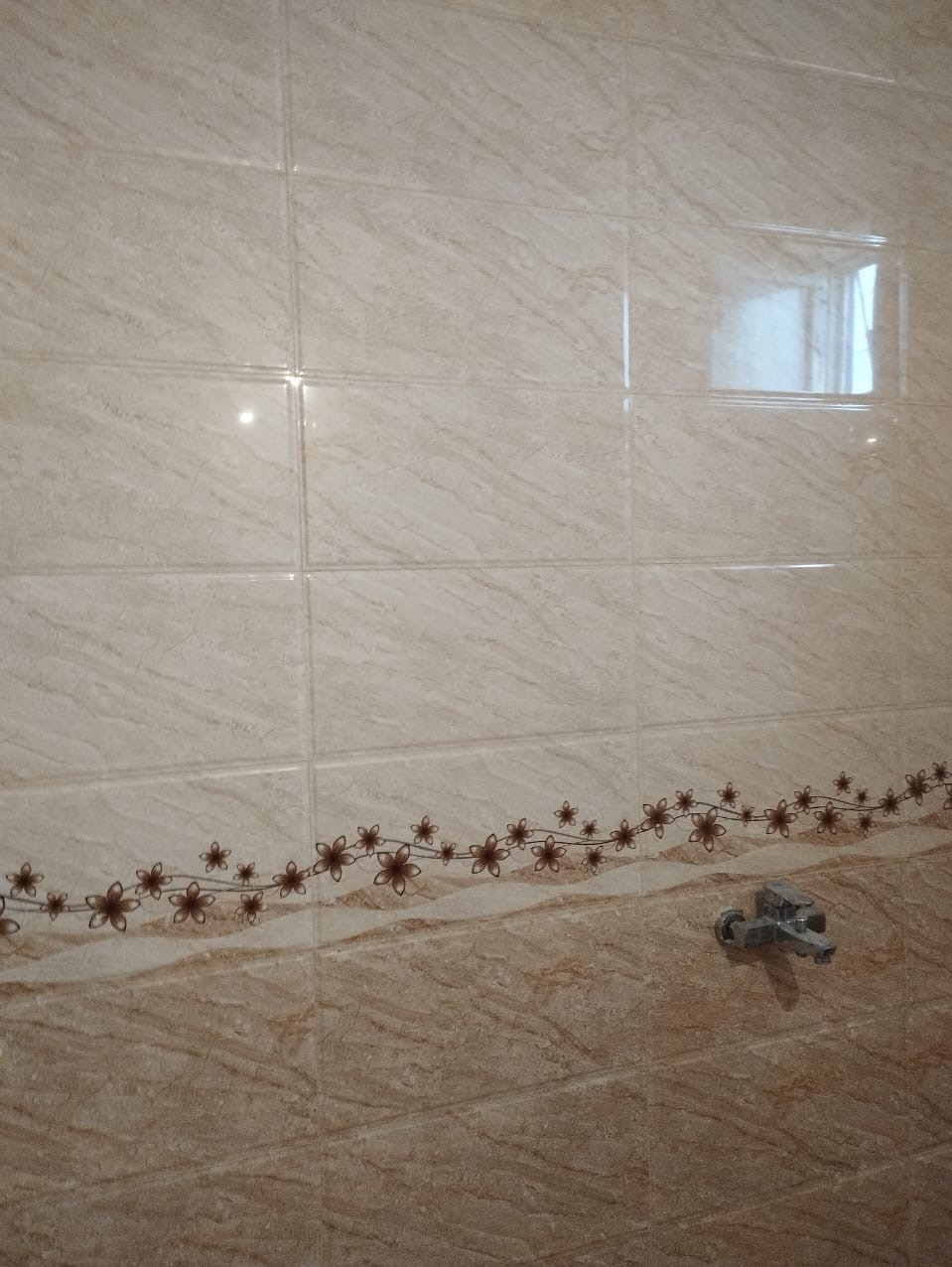 Beżowe płytki ceramiczne imitujące marmur z dekoracyjnym pasem kwiatowym i zamontowaną baterią umywalkową.