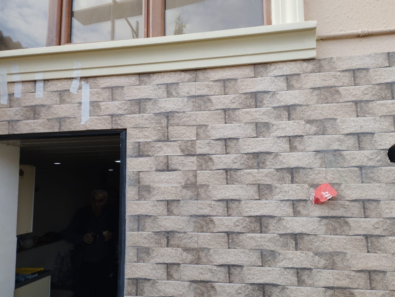Elewacja budynku wykończona płytkami imitującymi cegłę, z oknem w górnej części i wejściem widocznym po lewej stronie, fragment taśmy maskującej i czerwona naklejka na powierzchni płytek.