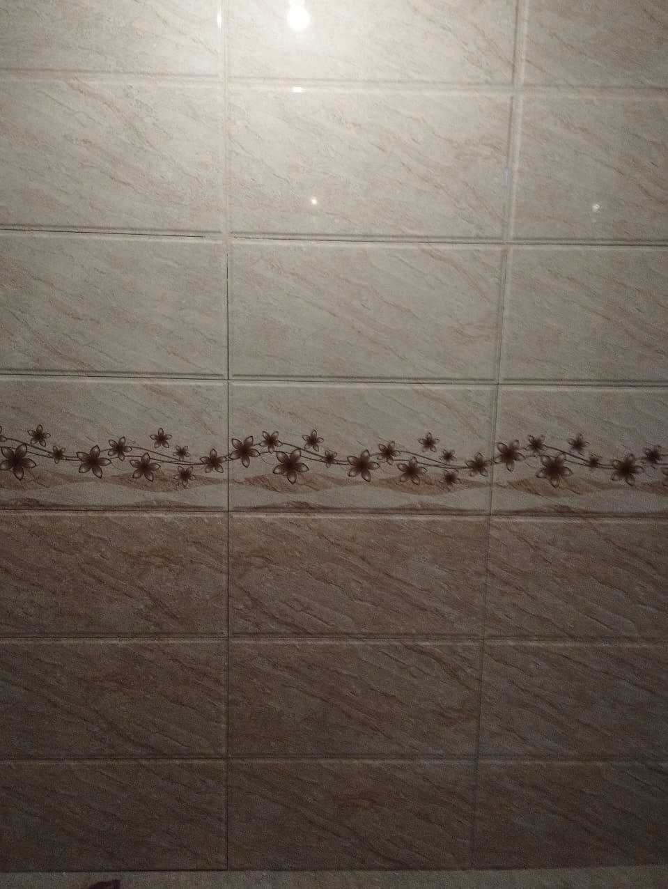 Ściana wyłożona beżowymi płytkami z delikatnym wzorem marmuru i poziomym pasem kwiatowym na środku.