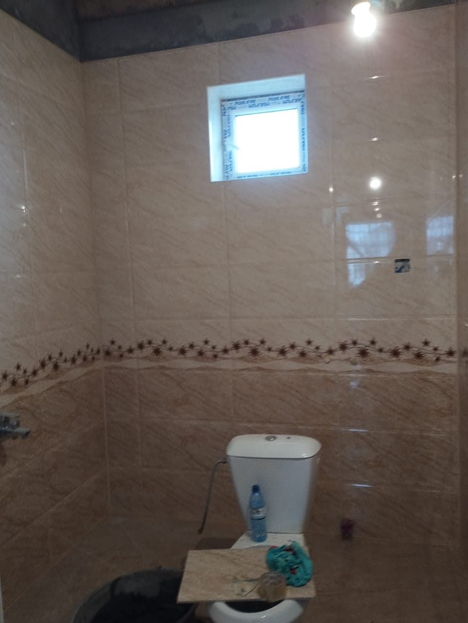 Wykończona łazienka z beżowymi płytkami, dekoracyjnym pasem z motywem kwiatowym, białą toaletą z deską tymczasowo przykrytą płytką i butelką wody na spłuczce, małe okno w górnej części ściany.