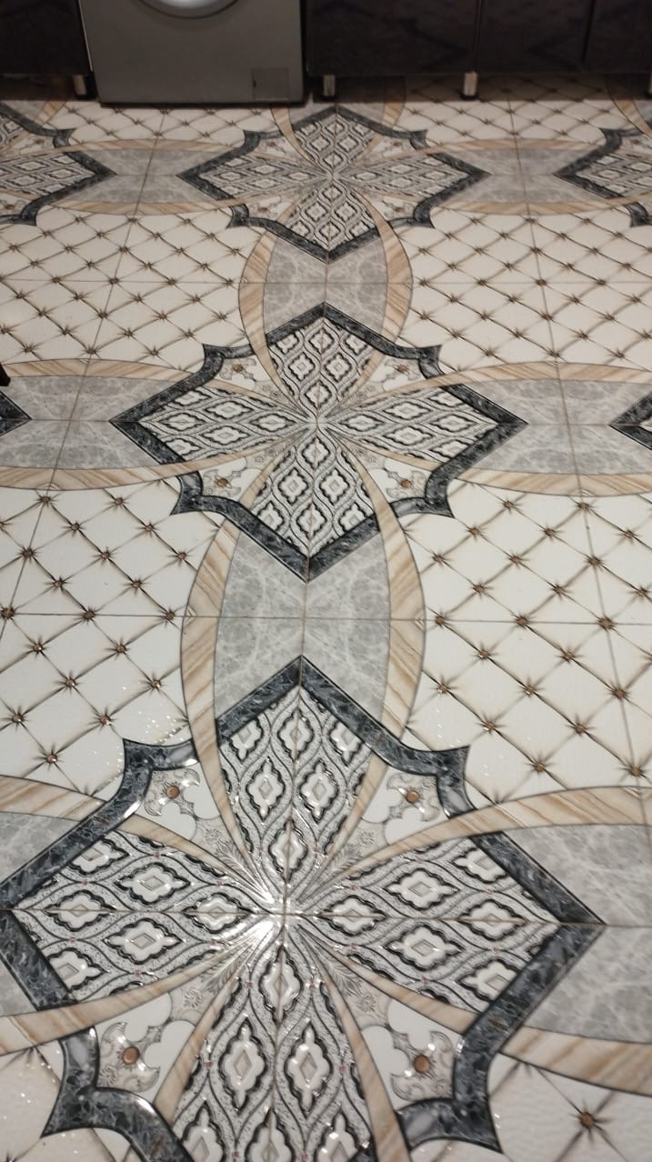 Wzorzysta podłoga z płytek ceramicznych w odcieniach bieli, szarości i beżu, z motywem przypominającym rozetę, widoczna z góry, z fragmentem białej szafki w tle.