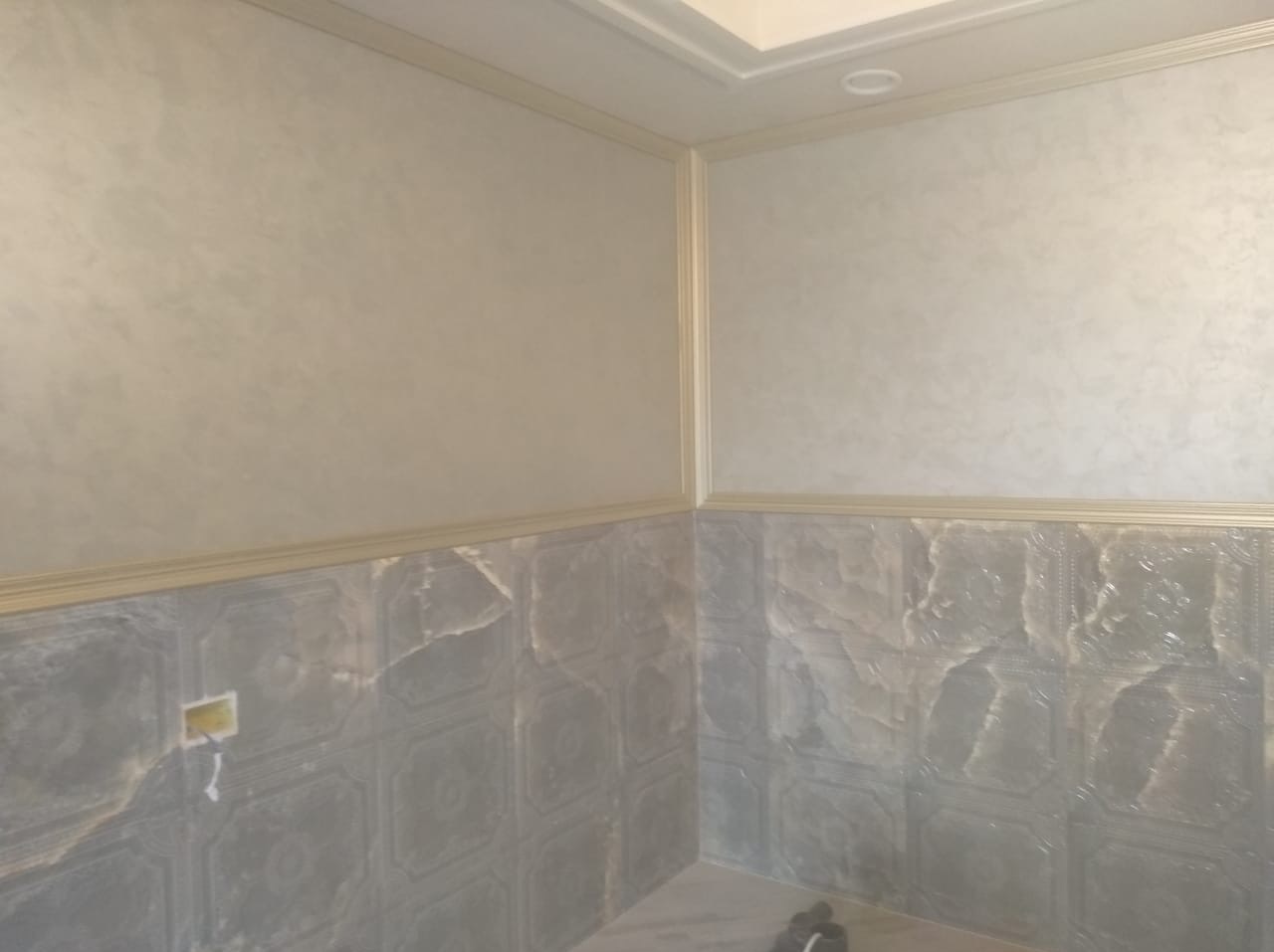 Wykończona łazienka z szarymi płytkami imitującymi marmur na dole ścian i beżową, teksturowaną farbą na górze, oddzielone ozdobną listwą. Widoczny fragment sufitu z oświetleniem.