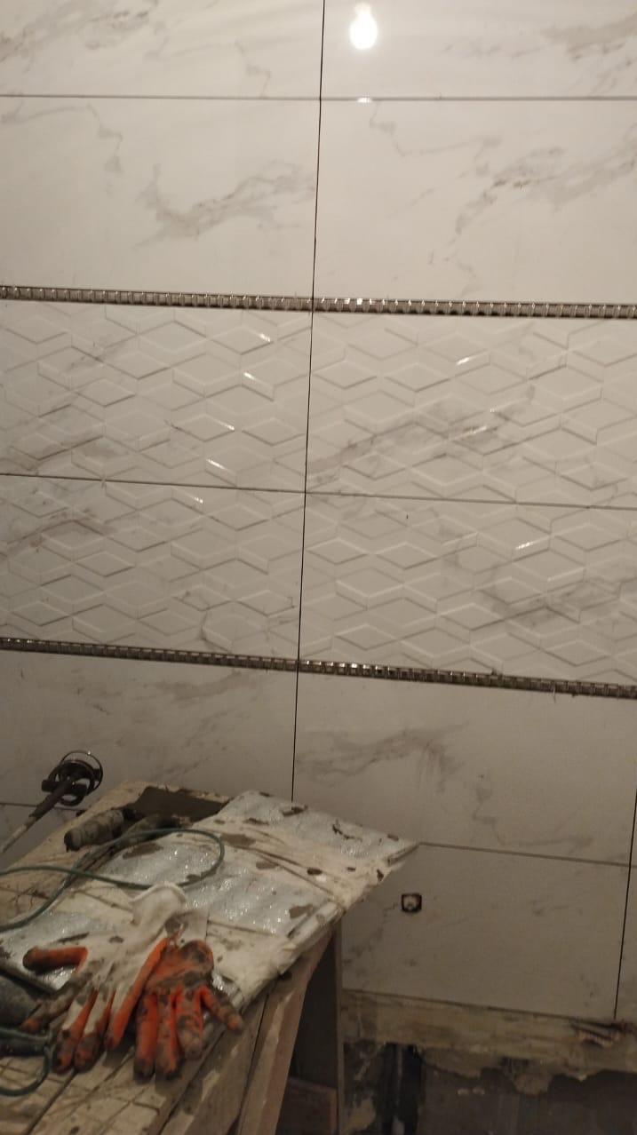 Układanie białych płytek z marmurowym wzorem i dekoracyjnym paskiem. Widoczne narzędzia i brudne rękawice na prowizorycznym stole.