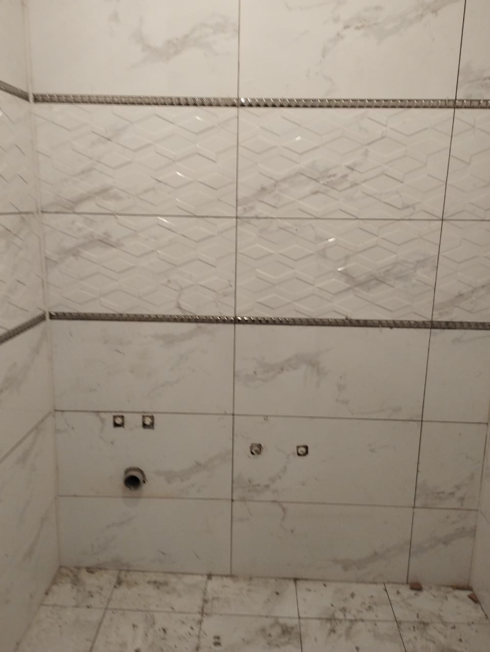 Ściana łazienki wyłożona białymi płytkami imitującymi marmur z ozdobną listwą, widoczne otwory na armaturę i częściowo ułożona podłoga z podobnych płytek.