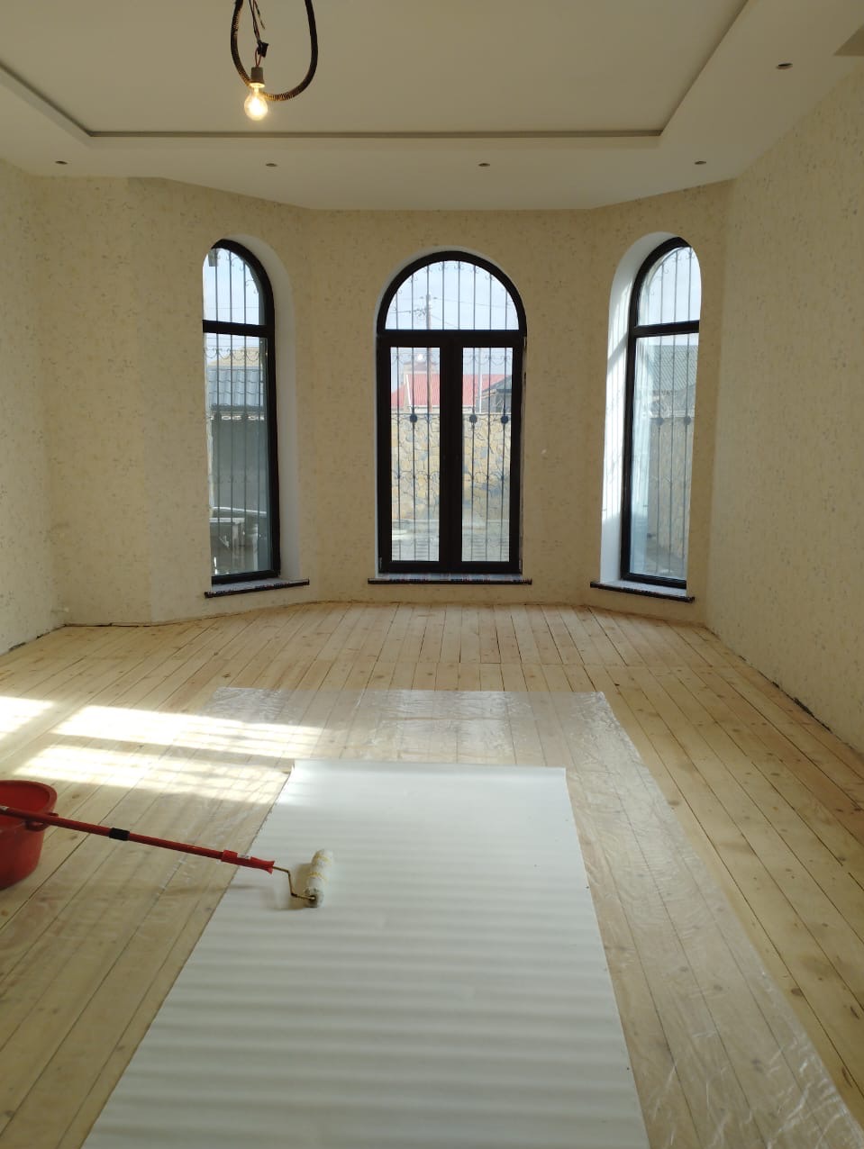 Przygotowanie podłogi drewnianej do malowania w jasnym pokoju z trzema oknami łukowymi, zabezpieczona folią ochronną i wałkiem malarskim.
