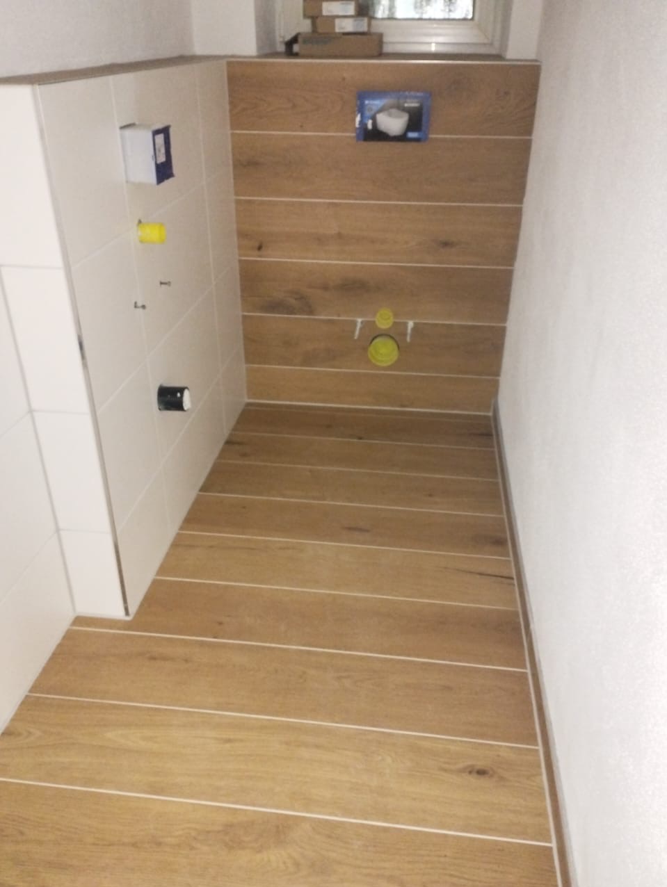 Przygotowanie do montażu podtynkowego stelaża WC na ścianie wyłożonej panelami drewnopodobnymi z widocznymi przyłączami, białe płytki na sąsiedniej ścianie, podłoga wyłożona panelami imitującymi...