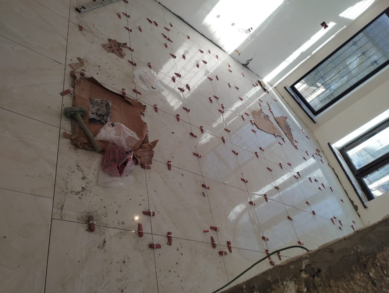 Układanie beżowych płytek podłogowych z użyciem czerwonych klipsów poziomujących, widoczne narzędzia glazurnicze i resztki opakowań na podłodze.