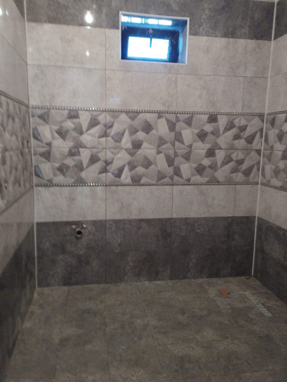 Wykończona łazienka z szarymi płytkami o różnych wzorach: geometrycznym na ścianie i jednolitym na podłodze, widoczny otwór wentylacyjny w górnej części ściany.