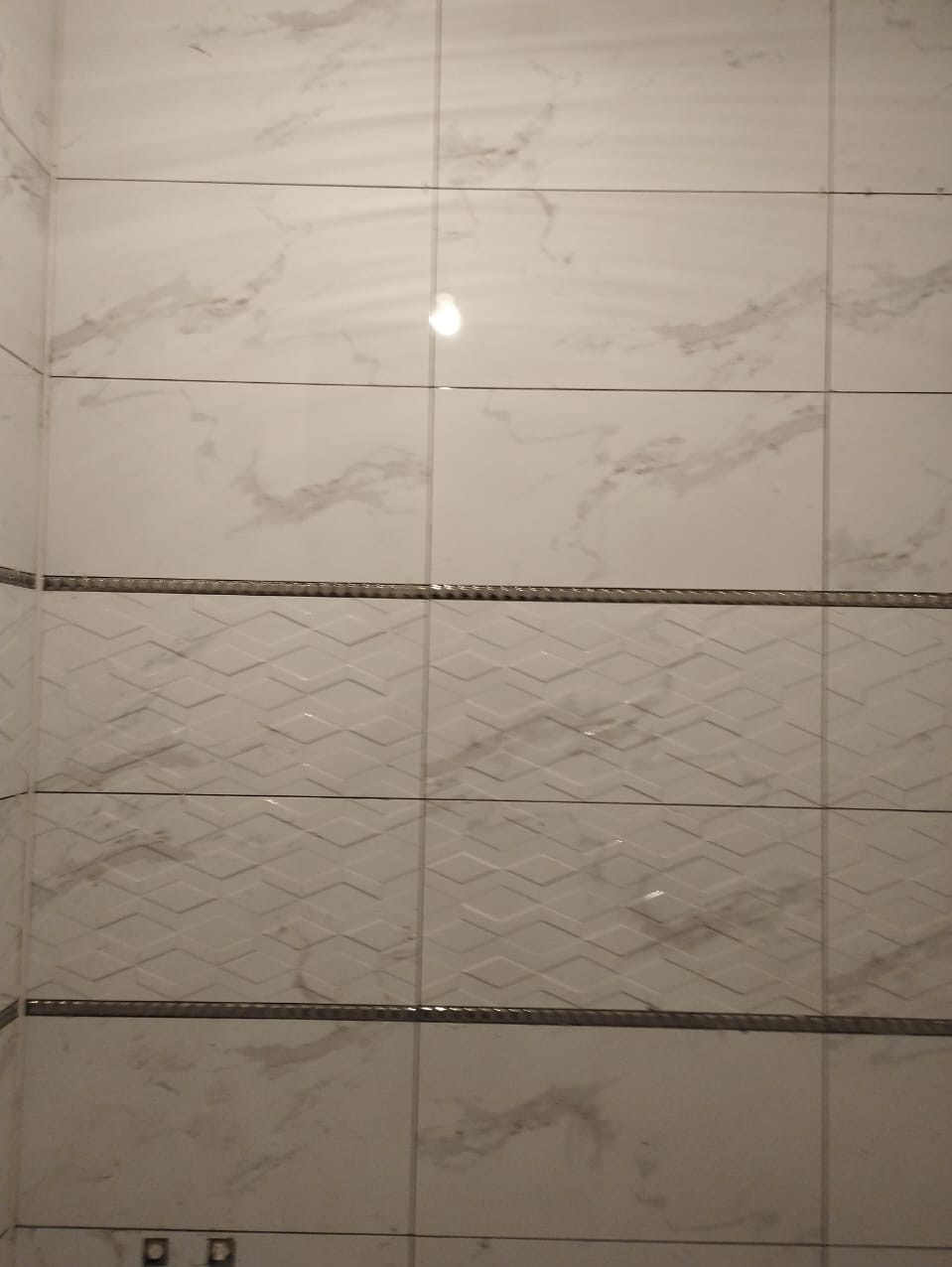 Wykończona ściana w łazience z białych płytek imitujących marmur, oddzielonych dekoracyjnym pasem z geometrycznym wzorem, widoczne fugi i odbicie światła.