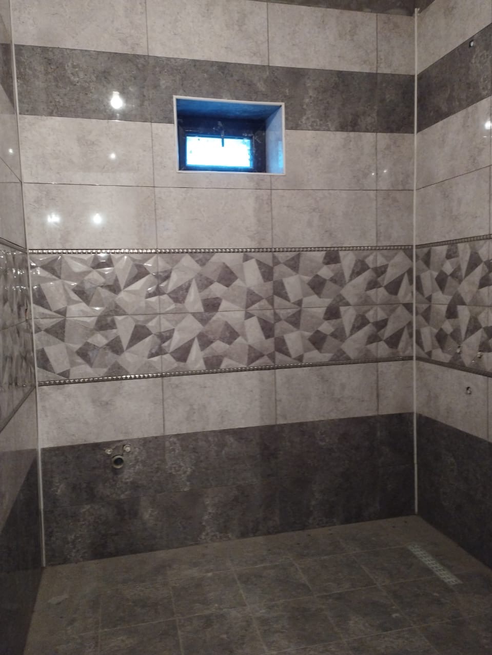 Wykończona łazienka z szarymi płytkami w różnych odcieniach i wzorach, małe okienko w górnej części ściany, widoczny odpływ liniowy w podłodze.