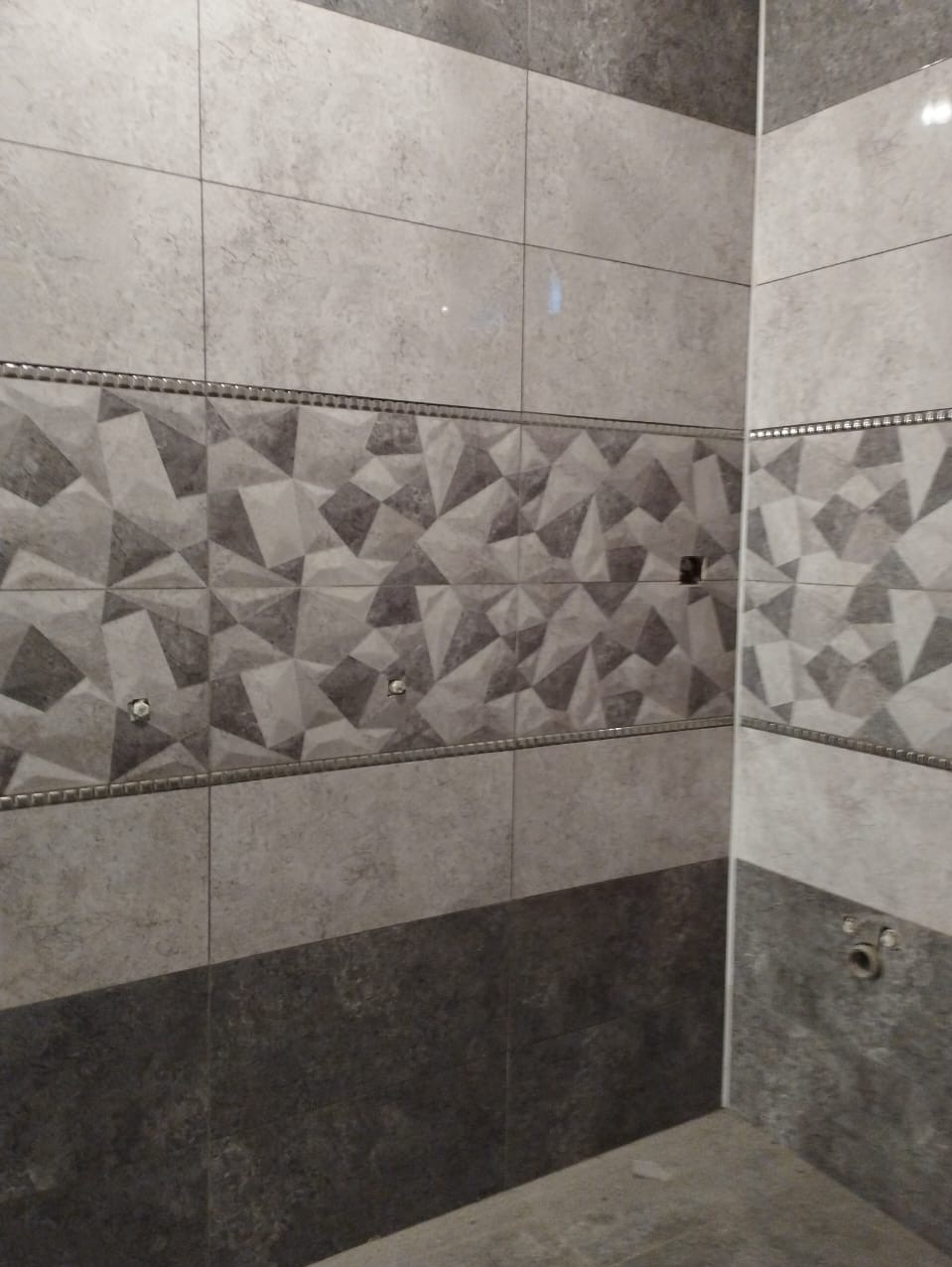 Ściana łazienki wyłożona szarymi płytkami z geometrycznym wzorem, widoczne otwory na armaturę i listwa dekoracyjna oddzielająca wzorzystą część od gładkiej.