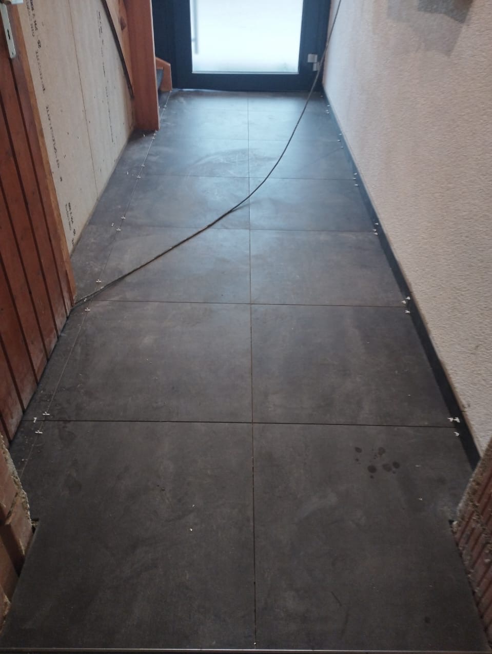 Układanie ciemnych, kwadratowych płytek podłogowych w korytarzu, widoczne krzyżyki dystansowe, przewód elektryczny i ślady po zabrudzeniach.