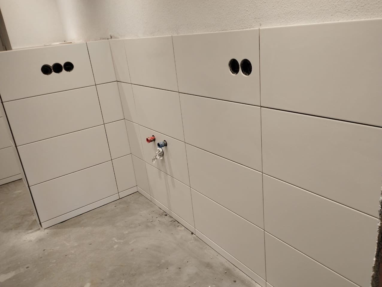 Ściana wyłożona białymi, prostokątnymi płytkami ceramicznymi z otworami na gniazdka elektryczne i wyprowadzeniem rur wodnych w trakcie prac wykończeniowych.