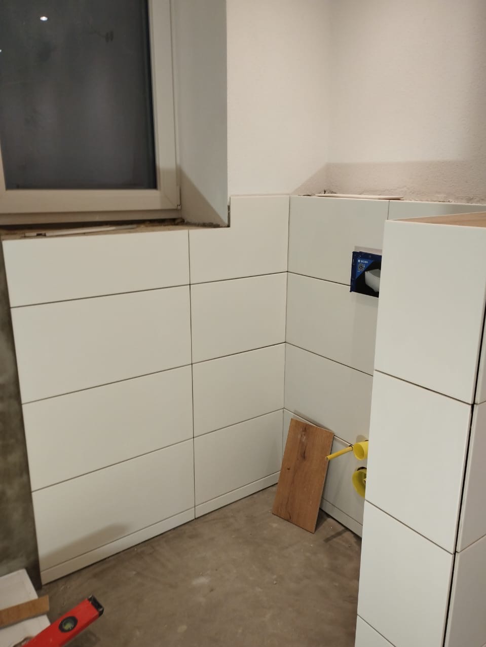 Świeżo ułożone, białe, prostokątne płytki ceramiczne na ścianie w rogu pomieszczenia, widoczne żółte rury instalacji wodnej i poziomica na podłodze.