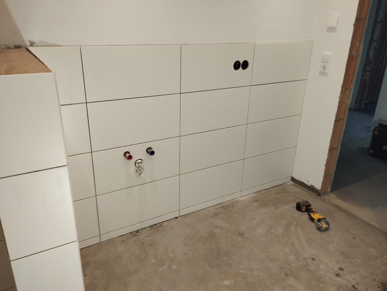 Świeżo położone białe, prostokątne płytki na ścianie, z widocznymi otworami na instalacje wodne i elektryczne, narzędzie do cięcia płytek leżące na podłodze.