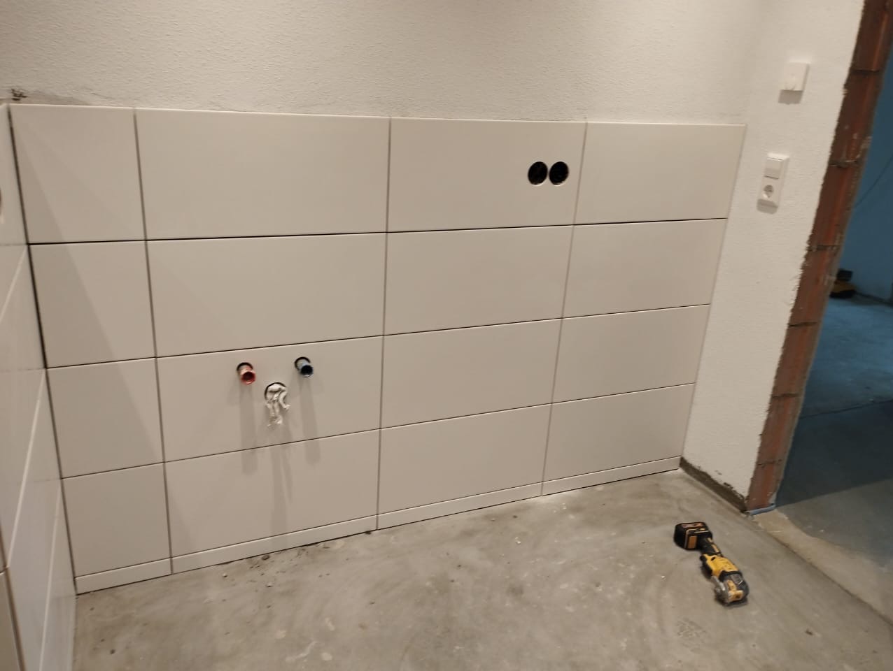 Ściana wyłożona białymi, prostokątnymi płytkami ceramicznymi, z widocznymi otworami na rury i gniazdka elektryczne, na podłodze leży narzędzie wielofunkcyjne.