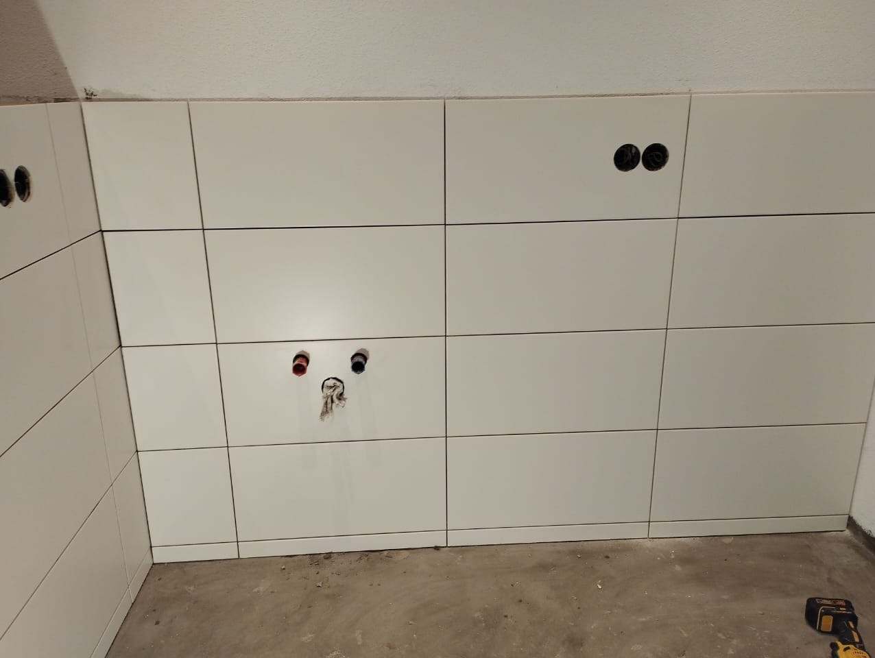 Ściana wyłożona białymi, kwadratowymi płytkami ceramicznymi, z widocznymi otworami na instalacje wodno-kanalizacyjne i elektryczne, oraz narzędzie akumulatorowe leżące na podłodze.