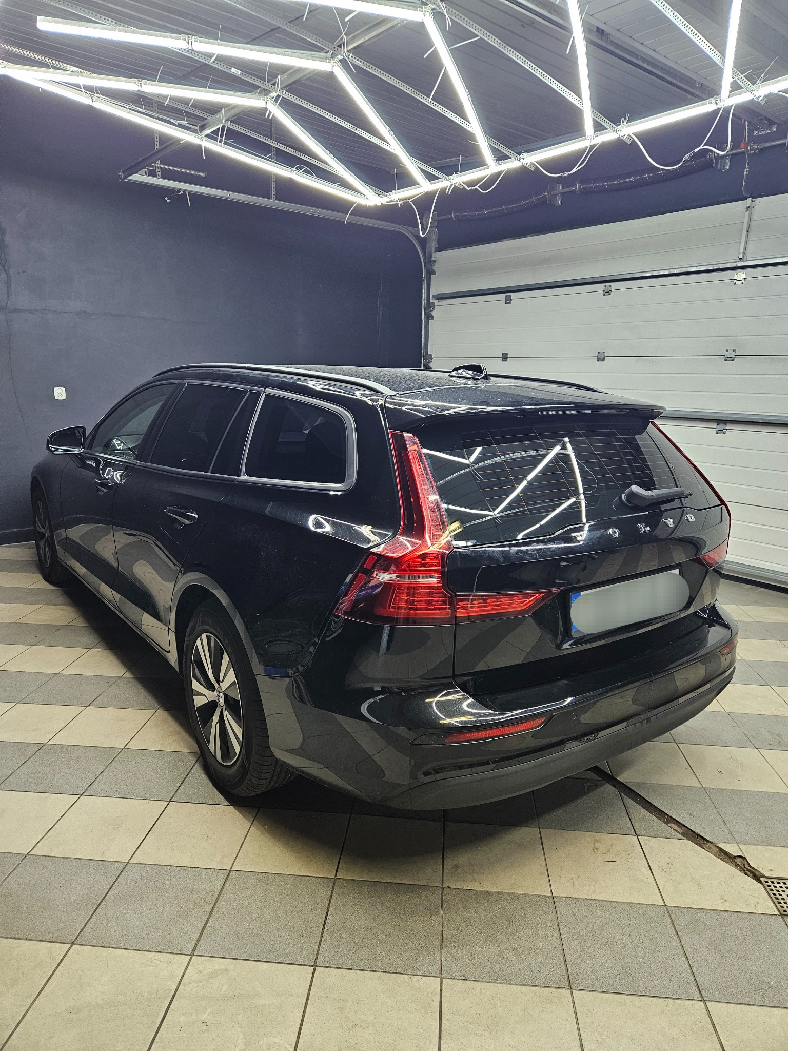 Czarny samochód kombi marki Volvo, widziany z tyłu, z przyciemnionymi szybami, stojący w garażu z jasnym oświetleniem i kafelkowaną podłogą.