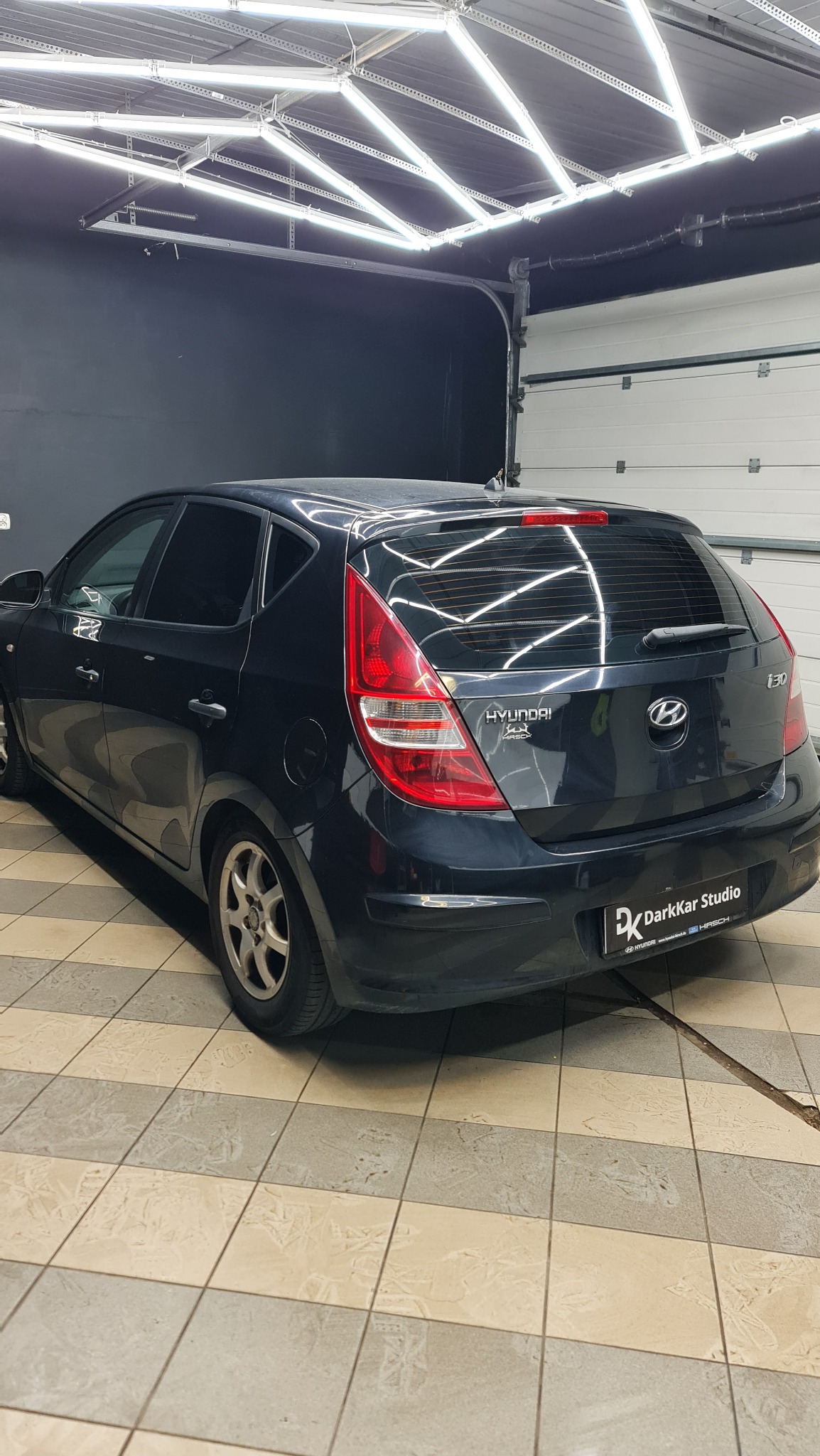 Ciemny Hyundai i30 z przyciemnionymi szybami, zaparkowany w garażu z jasnym oświetleniem sufitowym i białą bramą garażową po prawej stronie. Widoczny znak DarkKar Studio na tylnym zderzaku.